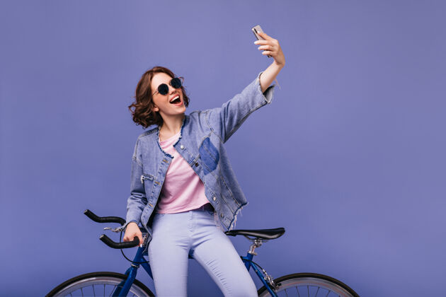 快乐梦幻般的白人女孩用手机和新自行车自拍漂亮的卷发女人戴着太阳镜站在自行车旁边自行车活跃骑行