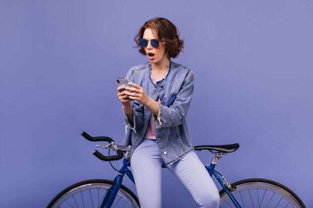 运动震惊的棕色头发的年轻女子看着手机屏幕壮观的女模特坐在自行车上 用她的手机骑手波浪发车辆