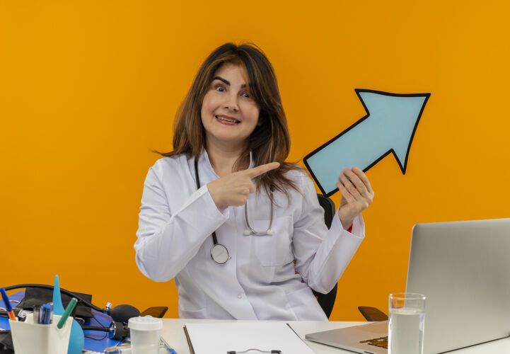 听诊器微笑的中年女医生穿着医用长袍和听诊器坐在办公桌旁 拿着医疗工具剪贴板和笔记本电脑 拿着箭头标记指着隔离的一侧微笑人中年