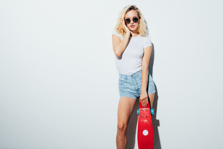 溜冰一个戴着太阳镜的快乐金发女人站在白色的墙上笑着和滑板合影的肖像可爱举行长度