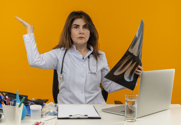 听诊器印象深刻的中年女医生穿着医用长袍和听诊器坐在办公桌旁 拿着医疗工具剪贴板和手提电脑 拿着x光照片 显示空手孤立女士笔记本电脑手