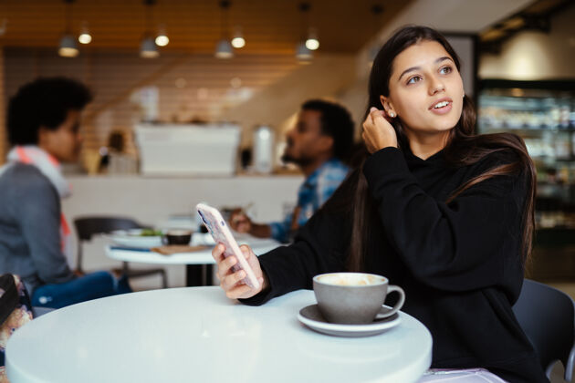 杯子年轻漂亮的女人用智能手机 迷人的女生坐在咖啡馆里肖像早餐女