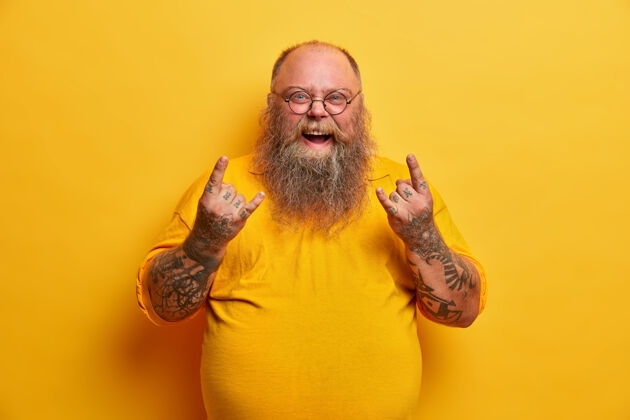 姿势穿着黄色t恤的肥胖搞笑男人 展示重金属标志 参加最喜欢的乐队的音乐会 大肚子 纹身手臂和胡须 戴着圆眼镜超重的摇滚迷在室内做手势体重休闲胃