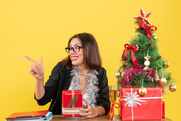 漂亮一位穿着西装 戴着眼镜 展示礼物的商务女士惊讶地指着右边的某个东西 坐在一张桌子旁 桌子上放着一棵圣诞树桌子眼镜指向