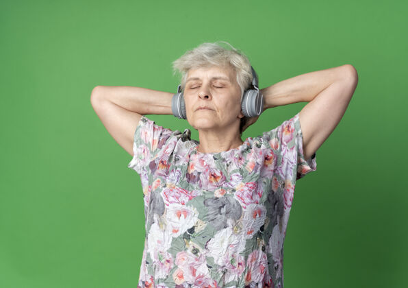 自信戴着耳机的自信的老妇人站在头后 双手紧握 闭着眼睛孤立地站在绿色的墙上站着抱着眼