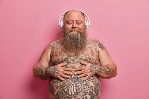 穿快乐快乐的大胡子男人双手放在大肚子上 戴着高质量的无线耳机欣赏美妙的音乐 有超重的问题 闭上眼睛 想象一些非常愉快的事情体重胃超重