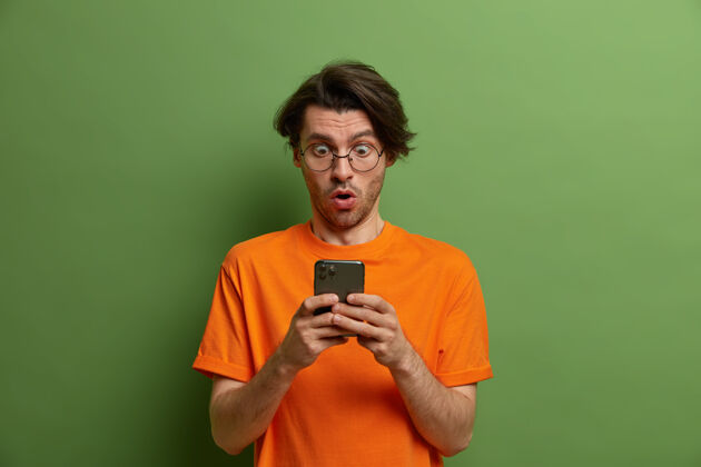 短信令人吃惊的男人的肖像凝视着智能手机屏幕 不敢相信自己的眼睛 得到令人震惊的信息 张开嘴屏住呼吸 穿着橙色t恤 对着绿色的墙壁摆姿势无语吃惊害怕