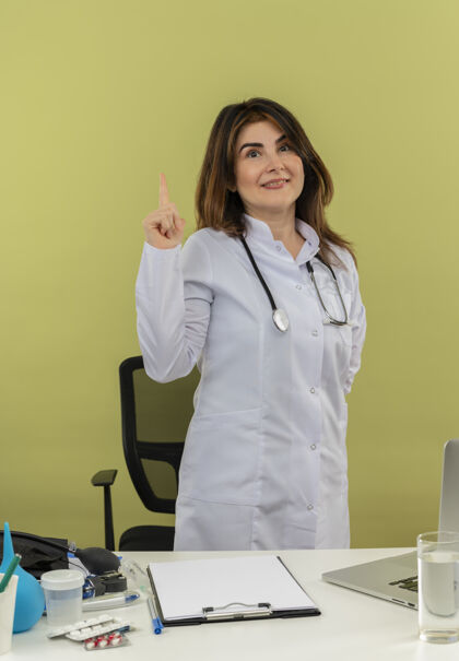 背部微笑的中年女医生穿着医用长袍和听诊器 站在桌子后面 拿着医疗工具和笔记本电脑 举起手指 把手放在背后隔离站立笔记本电脑工具