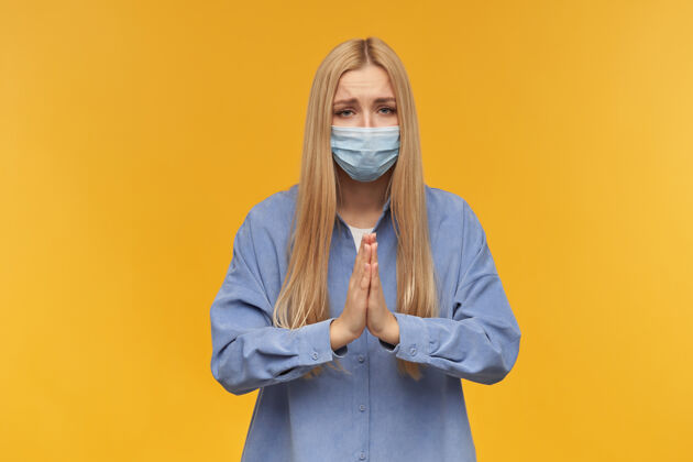 青少年十几岁的女孩 快乐的金发女人穿着蓝色衬衫和医用面罩 祈祷人和情感概念看着 橙色背景下孤立衬衫漂亮脸