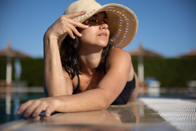 享受在阳光明媚的日子里 戴着草帽在游泳池附近的阳光下放松的迷人年轻女子度假和夏日概念阳光特写炎热