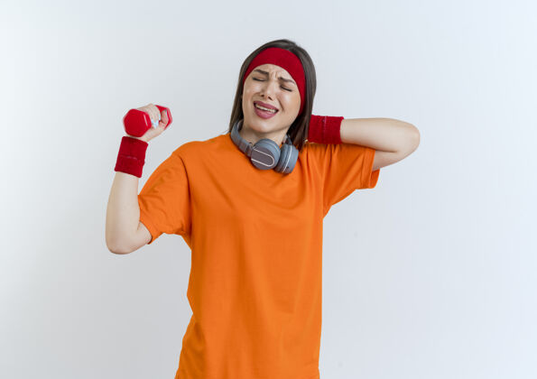 抱着疼痛的年轻运动女性戴着头带和腕带 脖子上戴着耳机拿着哑铃把手放在脖子后面闭着眼睛手运动年轻人