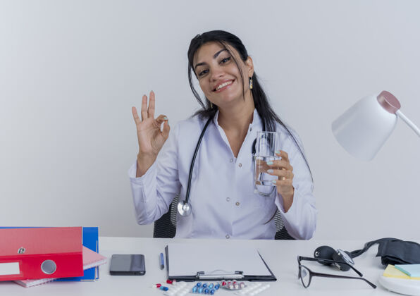 听诊器面带微笑的年轻女医生穿着医用长袍和听诊器坐在办公桌前拿着医疗工具望着手里拿着一杯水做着ok标志隔离可以年轻坐着
