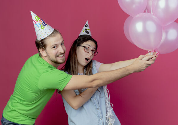 穿快乐的年轻夫妇戴着派对帽 手里拿着氦气球 看起来孤立在粉红色的墙上气球派对粉色
