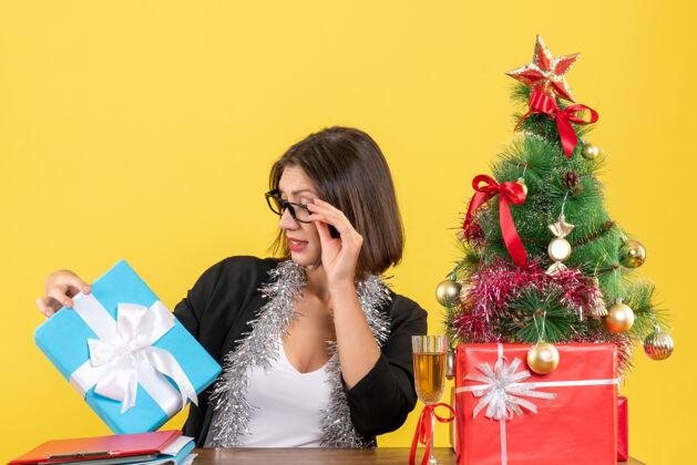 快乐情绪化的商务女士穿着西装 戴着眼镜 看着她的礼物 坐在一张桌子旁 桌子上放着一棵圣诞树微笑坐着眼镜
