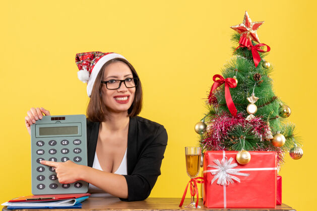 满意满意迷人的女士穿着西装 戴着圣诞老人的帽子和眼镜 在黄色的办公室里展示计算器漂亮西装显示