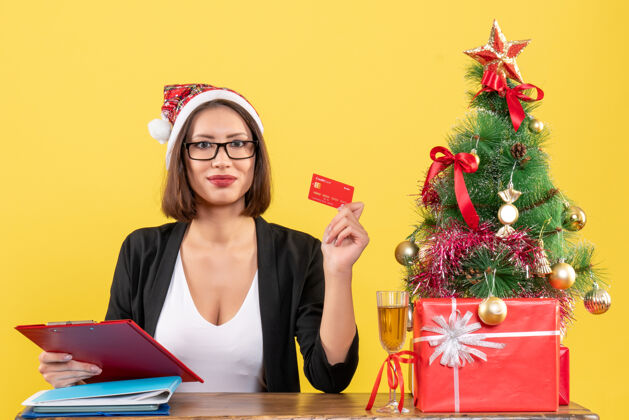 人正经迷人的女士穿着西装 戴着圣诞老人的帽子和眼镜 在黄色的办公室里出示银行卡圣诞老人成人漂亮