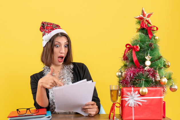 情绪情绪激动的商务女士穿着西装 戴着圣诞老人的帽子和新年装饰品 手里拿着文件 坐在一张桌子旁 桌子上放着圣诞树漂亮文件西装