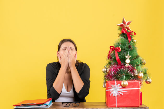 人穿着西装精疲力尽的年轻女子在黄色办公室装饰的圣诞树旁圣诞套装近