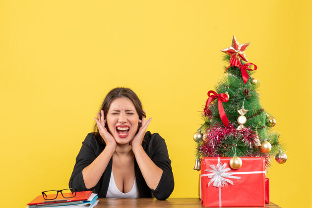 人情绪激动的年轻女子闭上耳朵坐在黄色办公室装饰圣诞树旁的桌子旁黄色年轻女子封闭