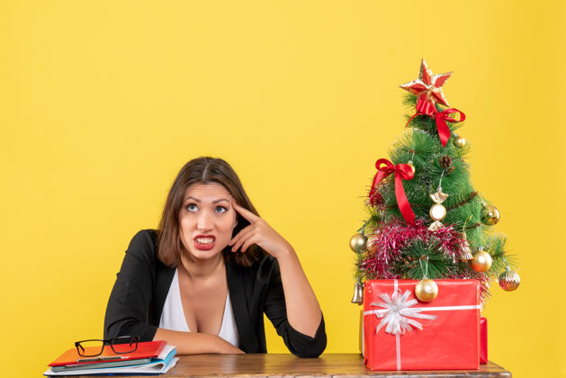 桌子困惑和紧张的年轻女子坐在一张桌子旁装饰圣诞树在黄色办公室紧张女人坐着
