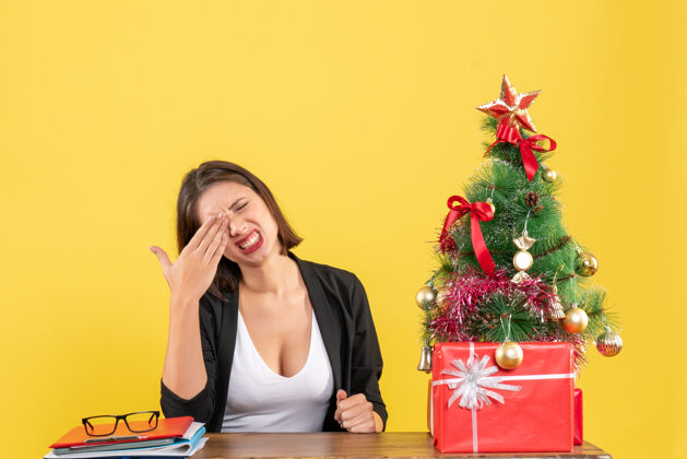 圣诞困惑的年轻女子穿着西装坐在一张桌子旁 在黄色的办公室装饰圣诞树旁快乐近树