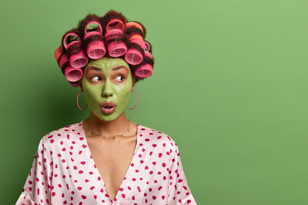 迷人令人印象深刻的震惊的女人为健康的皮肤使用新鲜的粘土面膜 做美容程序和面部护理 戴卷发器和家庭长袍 绿色墙壁上的空白惊艳表达尴尬