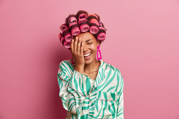 长袍搞笑快乐的女人做掌心 笑点什么 戴卷发 做完美的发型 穿睡衣 对着粉色的墙壁摆姿势女人 打扮和家居风格的概念乐观快乐肖像