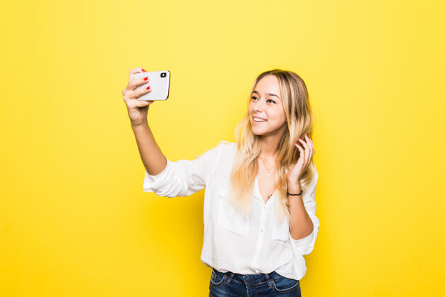 夏天照片中的女人手拿智能手机自拍隔离在黄色墙上时髦生动卷发