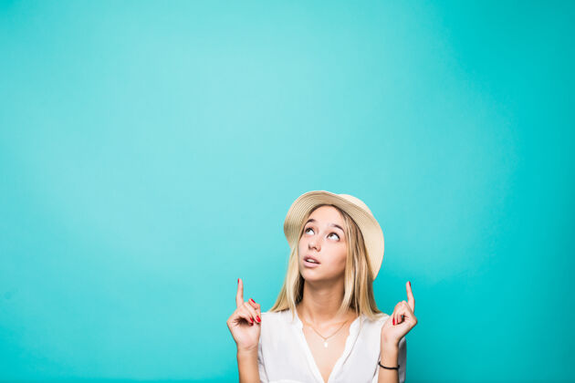 挑选一个戴着草帽 微笑着的美丽夏日女孩的肖像 两个手指指向蓝色墙壁上孤立的文字空间青年显示注意