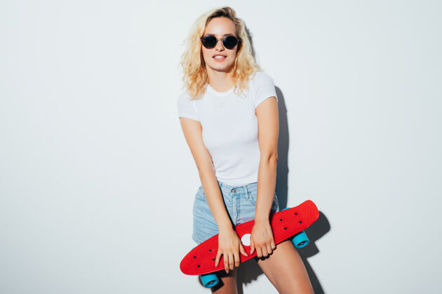 朋友一个戴着太阳镜的女人在白墙上举着滑板的肖像滑板乐趣微笑