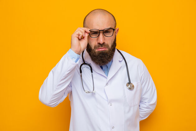 外套一位留着胡须的男医生 身穿白大褂 脖子上戴着听诊器 站在橙色背景上 透过眼镜近距离地看着摄像机眼镜支架周围