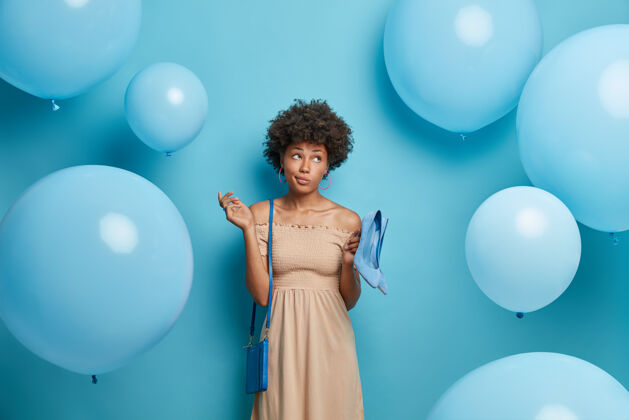 女人体贴的女人穿着米色长裙 拿着蓝色高跟鞋搭配包 在朋友周年纪念日到来之际 准备迎接节日活动 用充气气球隔着蓝色的墙壁尝试沉思年轻