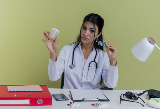 女士令人印象深刻的年轻女医生穿着医用长袍和听诊器坐在办公桌旁 手里拿着一包医用胶囊和烧杯工具拿着长袍