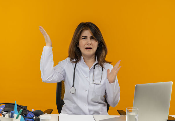 医生恼怒的中年女医生穿着医用长袍和听诊器坐在办公桌旁 手里拿着医疗工具 剪贴板和笔记本电脑 双手与空气隔绝笔记本电脑手橙色
