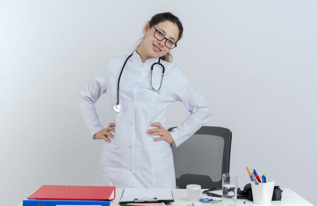 穿面带微笑的年轻女医生 穿着医用长袍 戴着听诊器和眼镜 站在桌子后面 手里拿着医疗工具 两手分开放在腰上医疗成年人医生