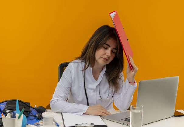 长袍疲惫的中年女医生穿着医用长袍和听诊器坐在办公桌前 用医疗工具 剪贴板和笔记本电脑触摸头部 用文件夹闭上眼睛隔离医生成人人