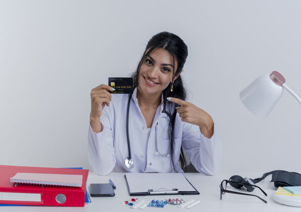听诊器面带微笑的年轻女医生穿着医用长袍 听诊器坐在办公桌旁 手里拿着医疗工具 看着信用卡指着它微笑成人医生