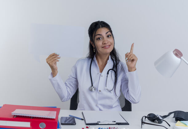 人令人印象深刻的年轻女医生穿着医用长袍和听诊器坐在办公桌旁 手持医疗工具 举起手指 看着孤立的聊天泡泡坐着印象抱着