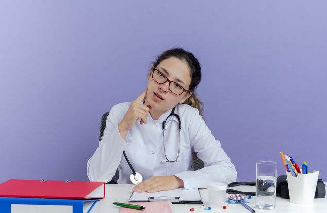 印象印象深刻的年轻女医生穿着医用长袍和听诊器坐在办公桌旁用医疗工具看着侧面触摸的脸穿着紫色触摸