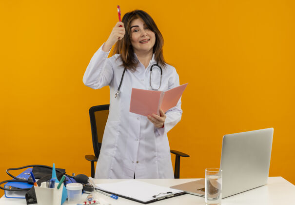 便笺微笑的中年女医生穿着医用长袍 听诊器站在桌子后面 带着医疗工具剪贴板和笔记本电脑 手里拿着笔和笔记本医生女性微笑