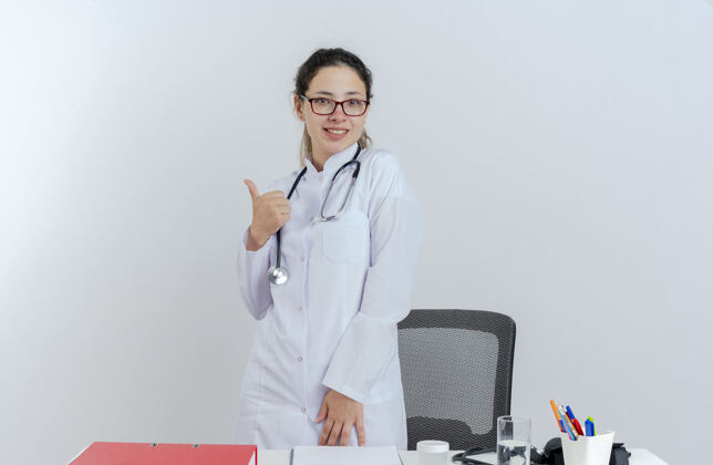 医生面带微笑的年轻女医生穿着医用长袍 戴着听诊器 戴着眼镜 站在桌子后面 拿着医疗工具 孤立地竖起大拇指桌子站立表演