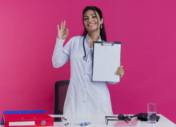 长袍面带微笑的年轻女医生穿着医用长袍 手持听诊器站在桌子后面 手持医疗工具 看着剪贴板 做着“ok”的手势粉色听诊器女士