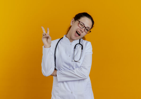 手势眨眼的年轻女医生穿着医用长袍 戴着眼镜听诊器 表现出和平姿态人显示长袍