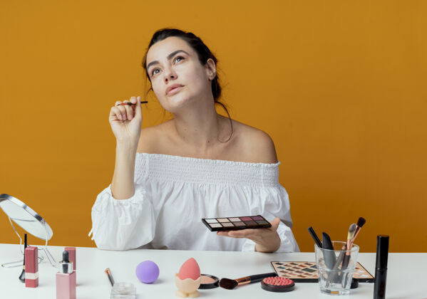 漂亮快乐美丽的女孩坐在桌子上用化妆工具拿着眼影调色板和化妆刷隔离在橙色墙壁上橘子调色板桌子