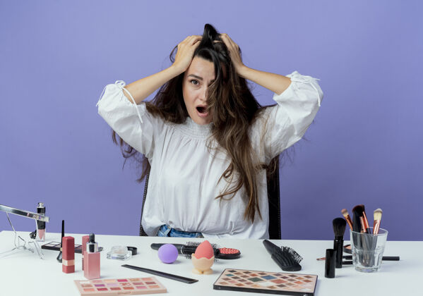 女人焦急的漂亮女孩坐在桌子旁 拿着化妆工具 把头抬在紫色的墙上 看起来很孤立紫色化妆焦虑