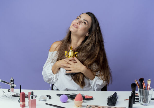 拿着高兴的漂亮女孩坐在桌子旁 拿着化妆工具 举着优胜者杯 孤零零地看着紫色的墙上向上美丽紫色
