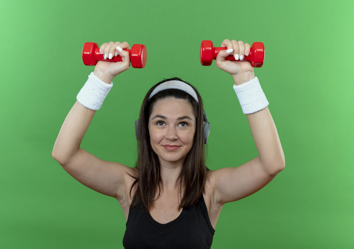 哑铃戴着耳机 戴着哑铃 自信地站在绿色背景下锻炼的年轻健身女士自信工作耳机