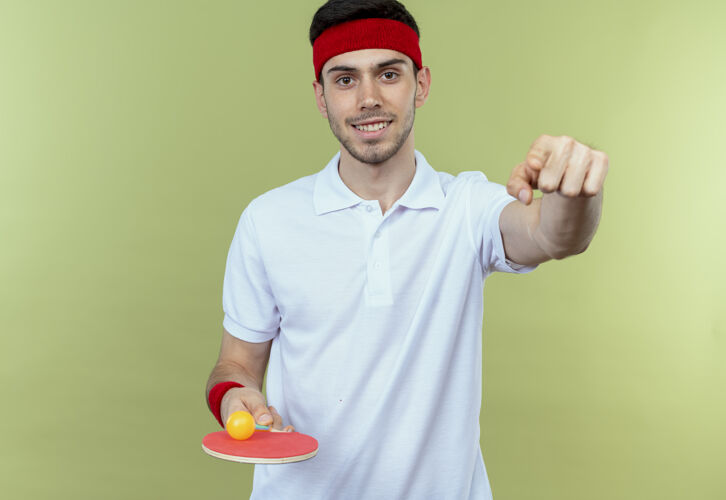 球拍戴着运动头巾的年轻人拿着球拍和乒乓球看着摄像机微笑着用食指指着站在绿色背景上的你球微笑桌子