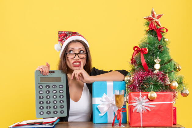 节目迷茫迷人的女士穿着西装 戴着圣诞老人的帽子 在办公室里展示着计算器上的黄色孤零零计算器黄色人