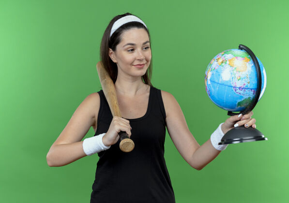 脸戴着头巾的年轻健身女士拿着棒球棒和地球仪站在绿色背景上面带微笑地看着它站着头带棒球
